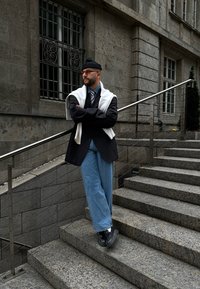 Άτομο φορώντας μαύρο σακάκι, ανοιχτό μπλε παντελόνι wide-leg και λευκή μπλούζα ριγμένη στους ώμους, στέκεται σε πέτρινα σκαλιά με μια κουπαστή.