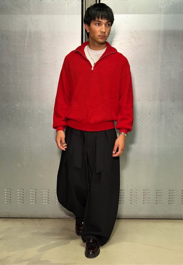 Person standing against a gray metal wall wearing a red pullover sweater, black wide-leg pants, dark shoes, and layered necklaces.
