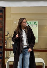 Hnedý semišový kabát cez károvanú košeľu a biele tričko, v kombinácii so svetlomodrými džínsami. Čierna taška dotvára outfit.