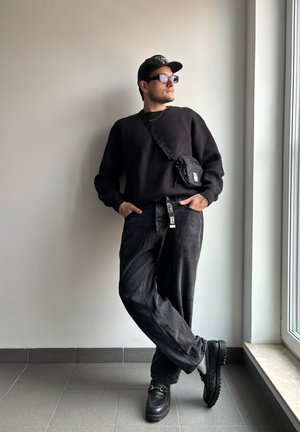 Suéter de punto negro, vaqueros negros de corte holgado, bolso cruzado negro, zapatos negros de suela gruesa y gorra negra con acentos de logo.