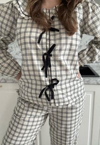 Femme debout dans la cuisine portant un pyjama en flanelle à carreaux crème et noir avec des rubans noirs noués sur le haut.