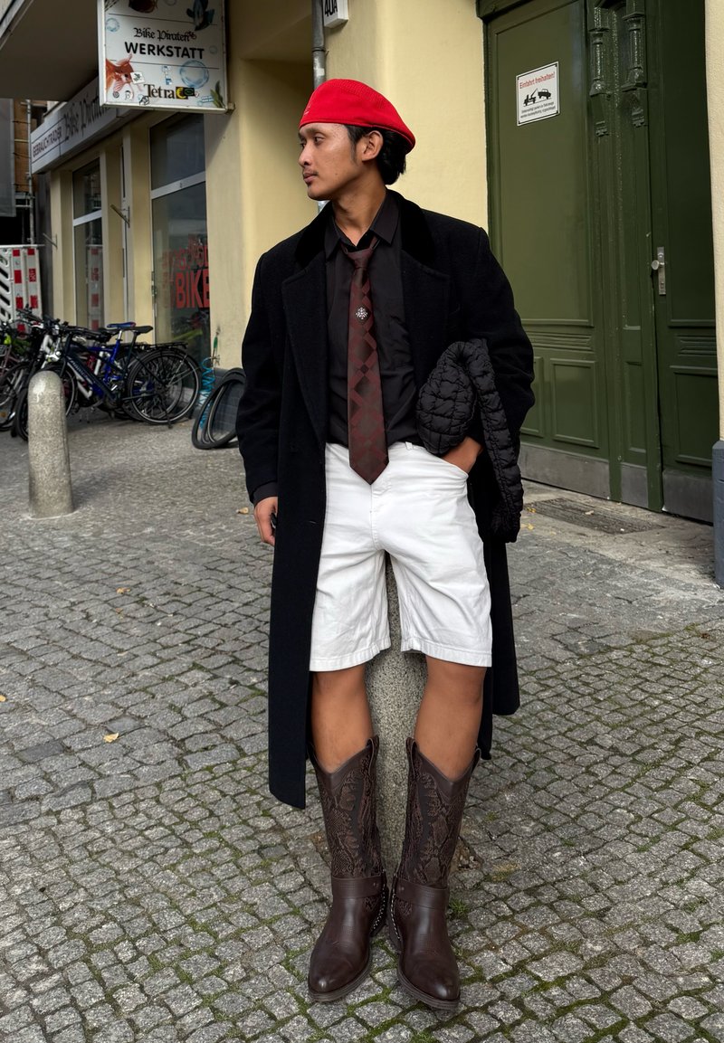 Black overcoat, black shirt with brown patterned tie, white shorts, and brown cowboy boots. Red beret and quilted black jacket in hand.