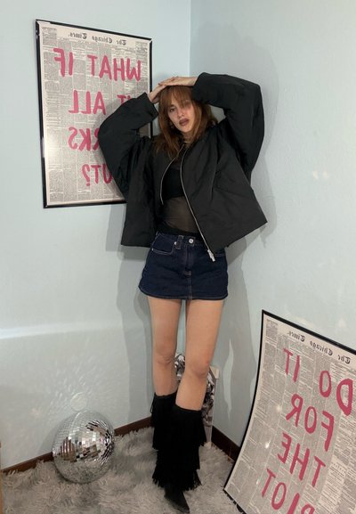 Joven mujer con chaqueta negra, falda vaquera y botas con flecos posando en una esquina con bola de discoteca y carteles de texto enmarcados en las paredes.