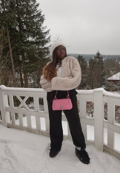 Chaqueta corta beige difusa, camisa blanca, pantalones de chándal negros, bolso rosa con herrajes plateados, de pie en la nieve cerca de una cerca de madera.