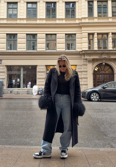 Abrigo negro largo con puños de peluche, camiseta negra y vaqueros azul claro. Zapatillas negras y blancas. Escenario urbano con edificios beige.