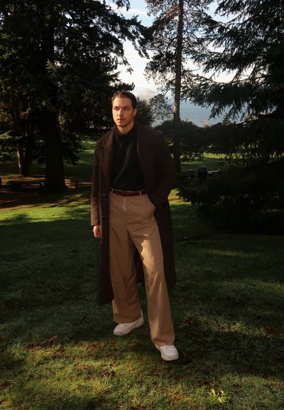 Hombre con abrigo largo oscuro, suéter negro y pantalones beige de pie sobre el césped en un parque con árboles y agua de fondo.