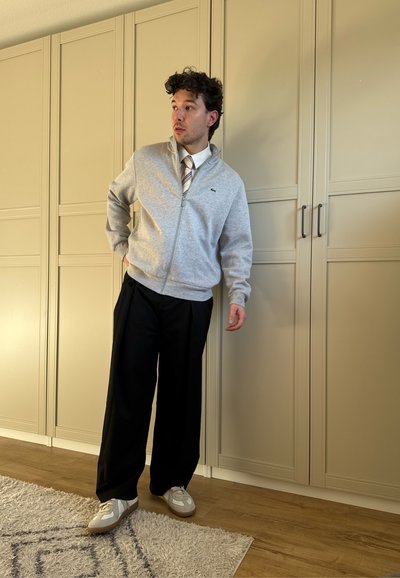 Joven con suéter gris con cremallera, corbata de rayas, pantalones anchos negros y zapatillas apoyado contra un armario beige en una habitación con suelo de madera.