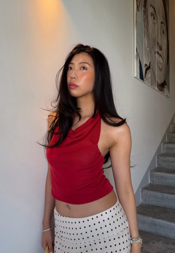 Jeune femme aux longs cheveux noirs porte un haut rouge sans manches et une jupe blanche à pois noirs, debout près d'un mur et d'un escalier à l'intérieur.