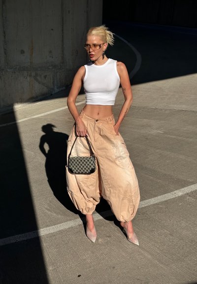 Top corto blanco, pantalones beige de gran tamaño con bolsillos, tacones altos transparentes y un pequeño bolso estampado. Fondo de concreto iluminado por el sol.