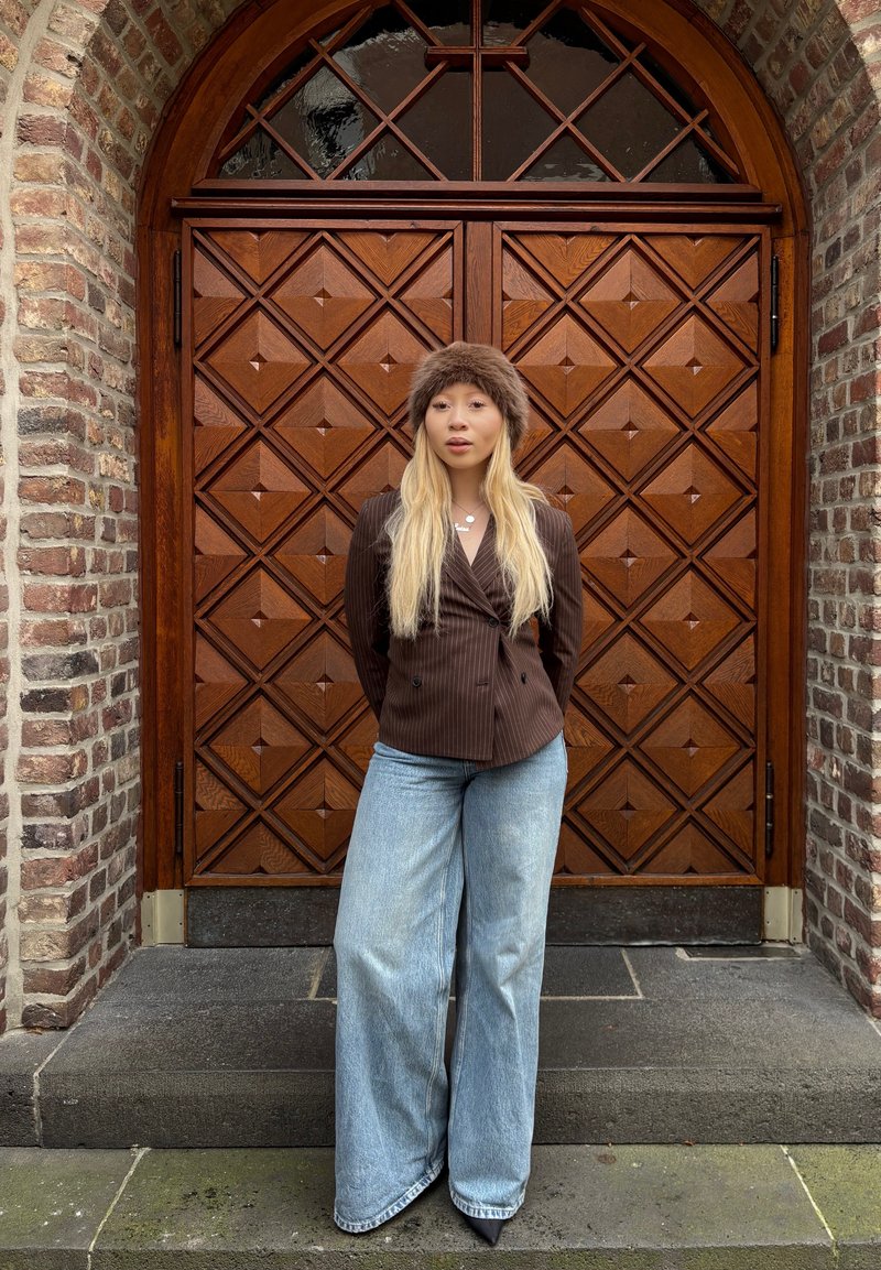 Hnědý pruhovaný dvouřadový blejzr, světle modré široké džíny a hnědý kožešinový klobouk, stojící proti texturovaným dřevěným dveřím s diamantovými vzory.