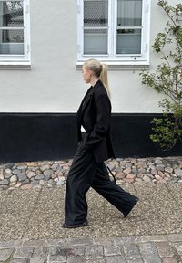 Zwarte blazer gecombineerd met losse zwarte broeken met studs. Model loopt over een kasseienpad naast een muur in neutrale kleuren.