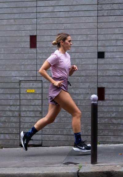 Camisa de manga corta rosa claro y pantalones cortos deportivos lavanda, zapatillas de correr negras y calcetines azules con estampado; corredor en movimiento frente a una pared texturada.