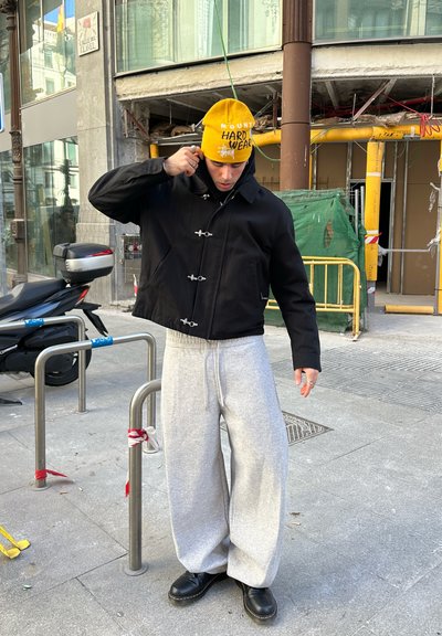 Chaqueta negra recortada con cierres de botones de palanca, pantalones deportivos gris de pierna ancha y un gorro amarillo con texto. Botas negras completan el conjunto.