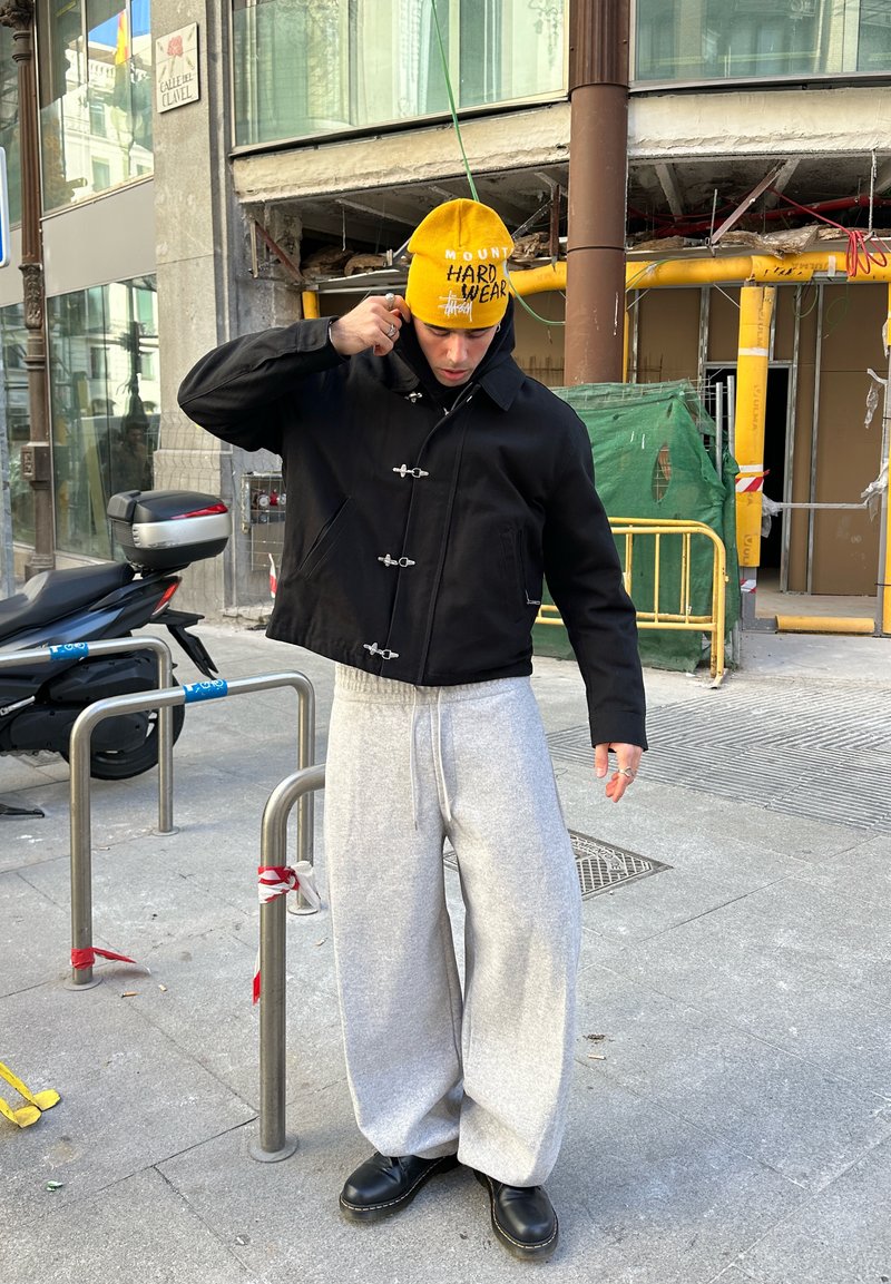 Black cropped jacket with toggle closures, grey wide-leg sweatpants, and a yellow beanie with text. Black boots complete the outfit.