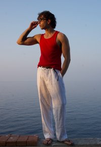 Top de alças vermelho com textura, calças de linho brancas e chinelos. Pessoa posa contra um fundo do oceano, com a mão a proteger os olhos da luz solar.