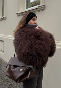 Manteau en fourrure marron duveteuse avec un col montant, associé à un pull gris foncé et un sac à main en cuir marron avec un accent argenté.