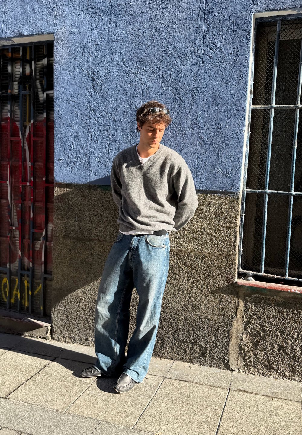 Pull gris en tricot, T-shirt blanc, jean bleu délavé ample et chaussures noires. En arrière-plan, un mur bleu texturé et une fenêtre à barreaux.