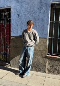 Pull gris en tricot, T-shirt blanc, jean bleu délavé ample et chaussures noires. En arrière-plan, un mur bleu texturé et une fenêtre à barreaux.