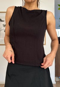 Čierny bezrukávový top z mäkkého materiálu, s obtiahnutým strihom a jemnými švami. Nosený cez čiernu sukňu. Pozadie obsahuje zarámované dielo.