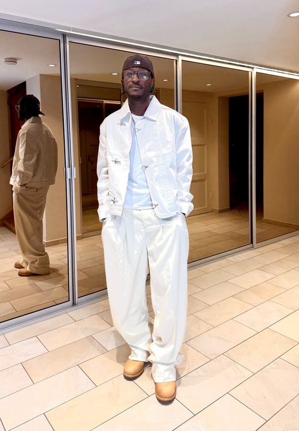 Uomo che indossa giacca e pantaloni bianchi, stivali color cuoio e un cappello scuro indossato al contrario, in piedi con le mani in tasca vicino a porte in vetro riflettente all’interno.