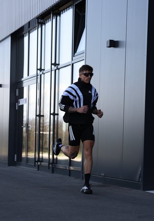 Hombre vestido con ropa deportiva negra y blanca y gafas de sol, corriendo delante de un edificio moderno de vidrio y metal.