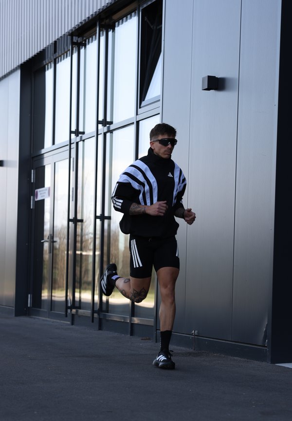 Mężczyzna ubrany w czarno-biały strój sportowy i okulary przeciwsłoneczne biegnie obok nowoczesnego budynku ze szkła i metalu.