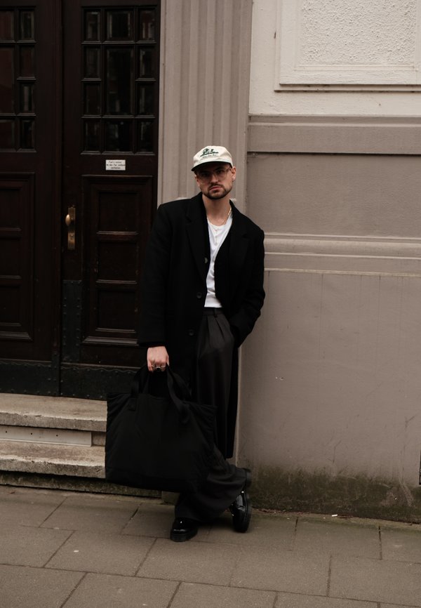 Musta pitkä takki valkoisen paidan, harmaiden housujen ja mustien kenkien päällä. Vaalea pipo ja iso musta laukku. Seisoo tekstuuriseinää vasten.