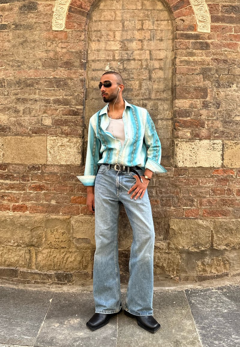 Homme portant des lunettes de soleil, une chemise à rayures turquoise, un débardeur blanc, un jean bleu clair et des bottes noires, debout contre un mur en brique texturé.
