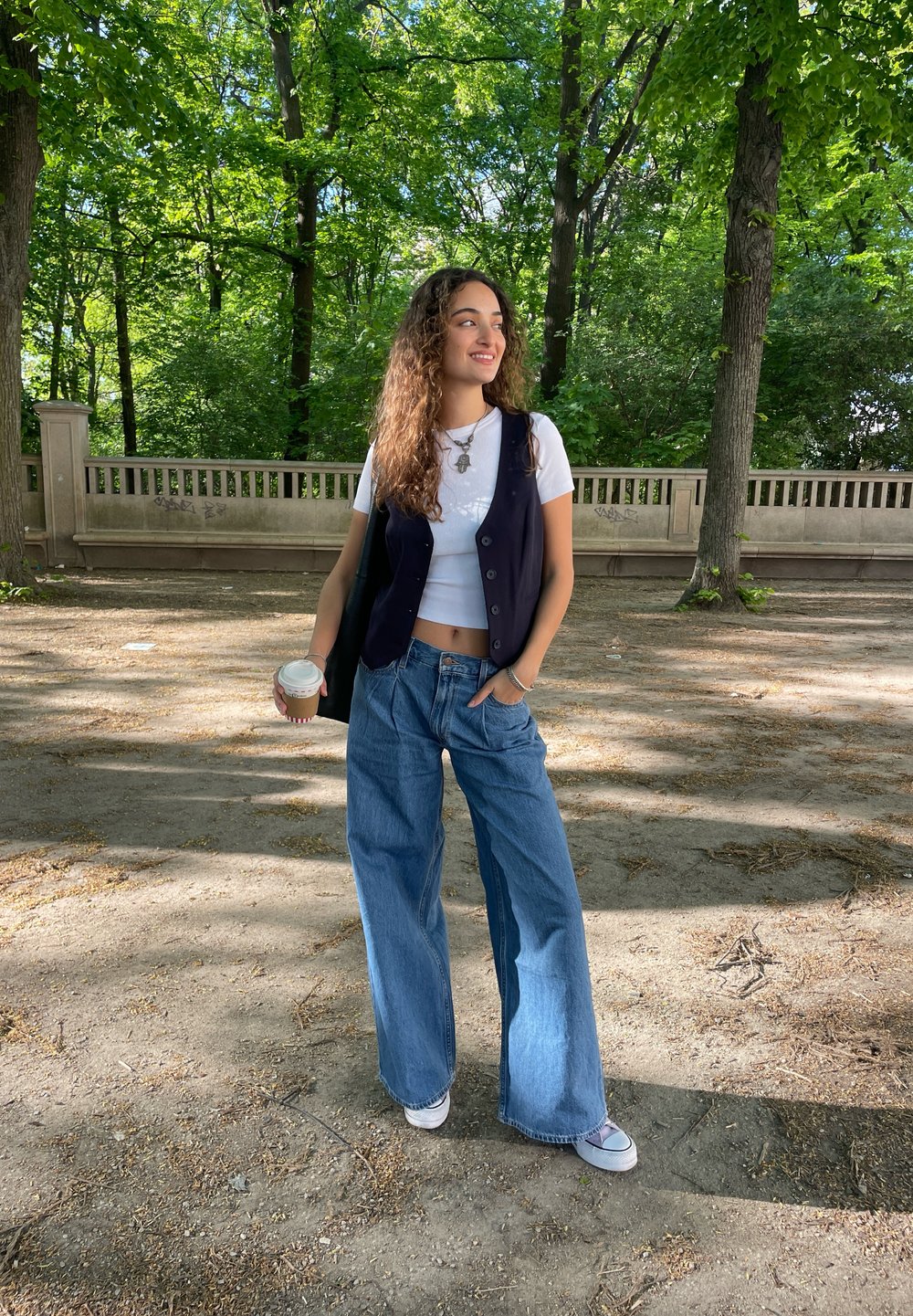 Μαύρο γιλέκο πάνω από ένα λευκό t-shirt, συνδυασμένο με ανοιχτό μπλε φαρδιά τζιν παντελόνια και λευκά αθλητικά παπούτσια. Κρατάει ένα ποτήρι καφέ και στέκει έξω.