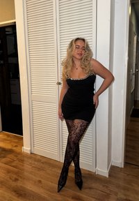 Femme aux longs cheveux blonds, en robe noire, collants en dentelle florale et talons, posant à l'intérieur devant des portes blanches de placard.