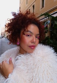 Femme aux cheveux bouclés portant un manteau blanc duveteux, des boucles d'oreilles roses et un maquillage des yeux rose vif, debout dans une rue de la ville.
