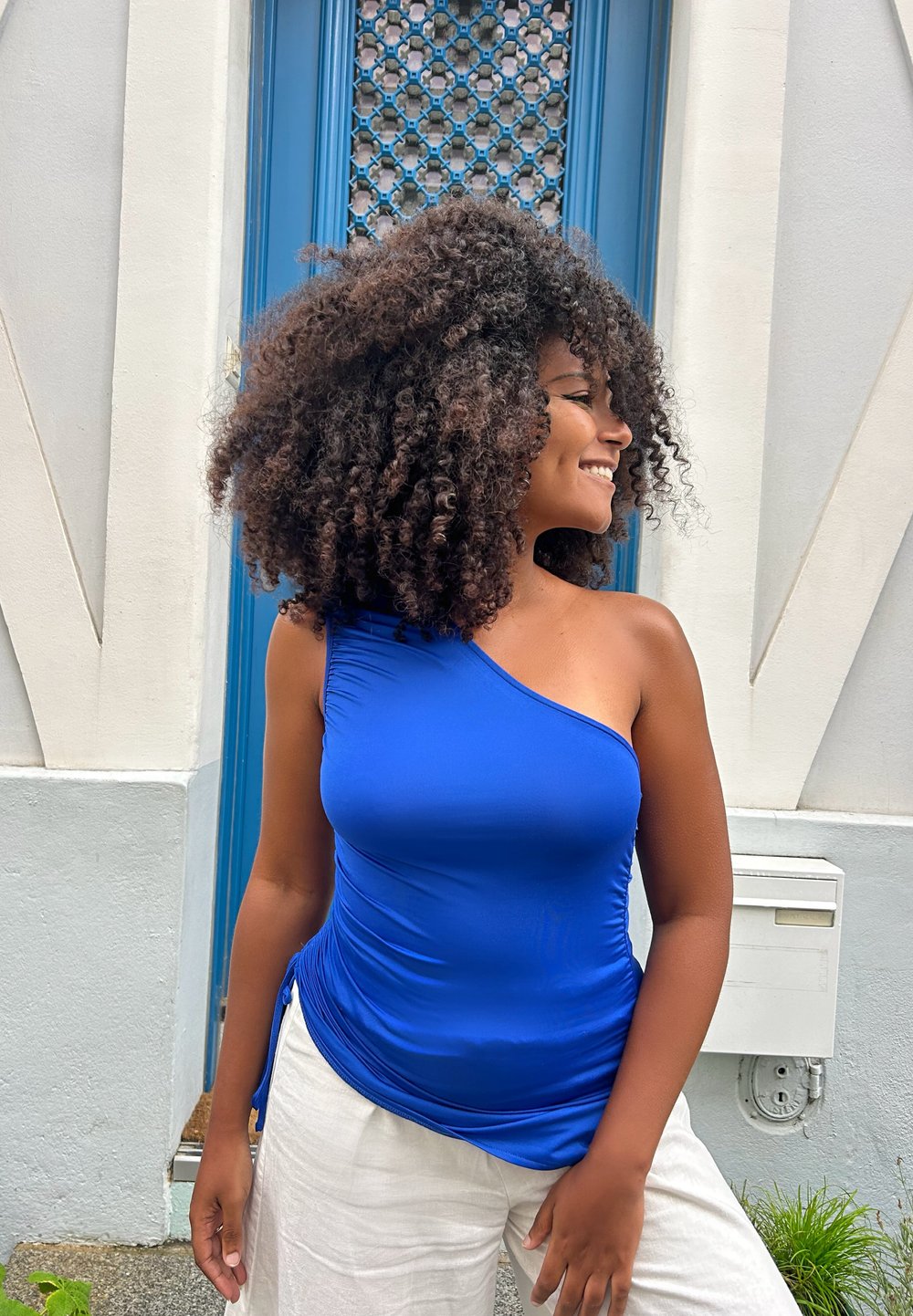 Top blu reale a spalla unica in tessuto elasticizzato con dettaglio drappeggiato in vita. Indossato con pantaloni a gamba larga di colore chiaro. Sfondo di muro testurizzato.