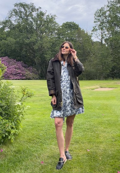 Mujer ajustándose las gafas de sol, con vestido estampado y chaqueta oscura, de pie sobre hierba verde con árboles y arbustos floridos en el fondo.