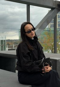 Casaco preto oversized com colarinho alto, combinado com uma saia preta. Os acessórios incluem óculos de sol estreitos e uma pequena mala preta.