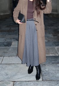 Brauner Wollmantel über einem dunklen Oberteil, grauer plissierter Rock und schwarze Knöchelstiefel. Die Accessoires umfassen eine kleine schwarze Clutch. Texturierte Stein-Hintergrund.