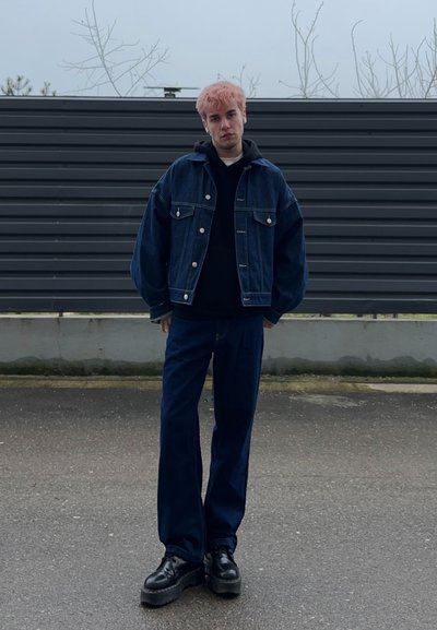 Joven con cabello rosado de pie sobre la acera, vistiendo una chaqueta vaquera oscura y oversize, sudadera negra con capucha, jeans anchos y zapatos plataforma negros.