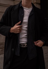 Melnā vilnas mētelis uz svītrainas krekla un baltas krekliņa, ar metāla jostas sprādzi un gredzeniem uz pirkstiem, izvietots pilsētvidē.