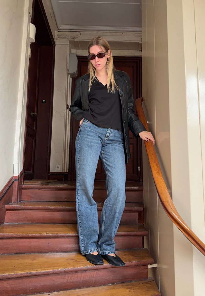 Femme portant des lunettes de soleil noires, une veste noire, une chemise noire, un jean bleu, debout sur des escaliers en bois tenant une rampe.