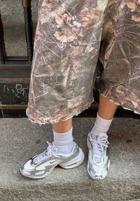Legs clad in white Nike trainers with silver details, paired with white ribbed crew socks and cropped camouflage trousers featuring a leaf pattern, standing on concrete.