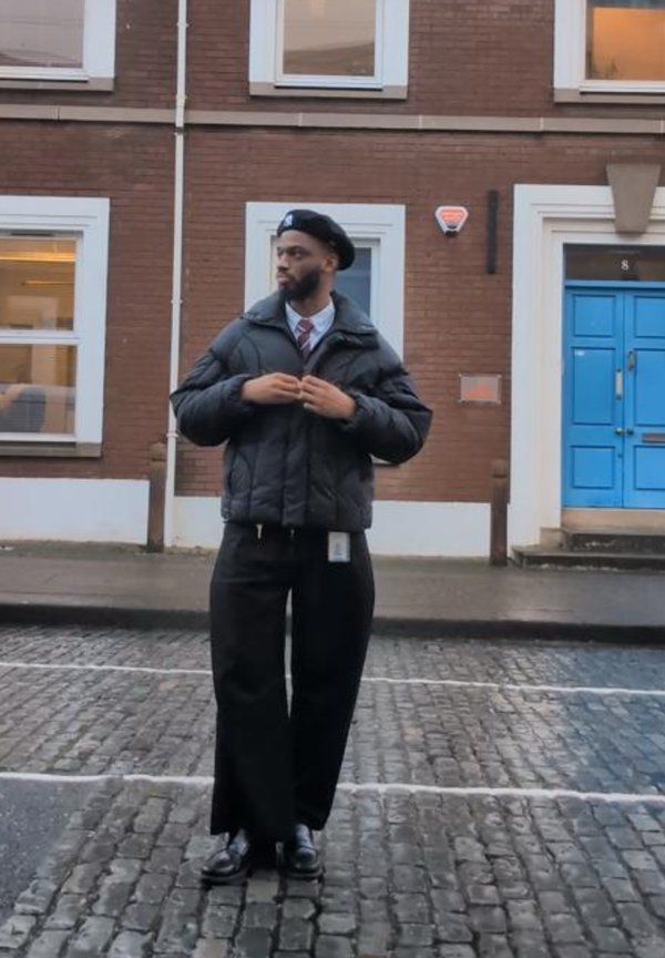 Mann mit schwarzer Baskenmütze, Steppjacke, weißem Hemd, Krawatte und weiten schwarzen Hosen, der auf einer Kopfsteinpflasterstraße neben einer blauen Tür steht.