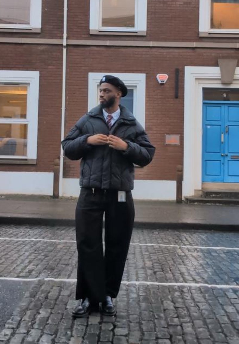 Homme portant un béret noir, une doudoune, une chemise blanche, une cravate et un pantalon large noir, debout sur une rue pavée près d'une porte bleue.