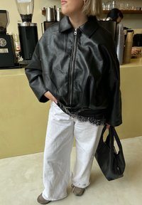 Veste en cuir noire avec fermeture éclair, coupe oversize, associée à un pantalon ample blanc. Un bordeau en dentelle noire est visible à la taille. Porte un sac noir.