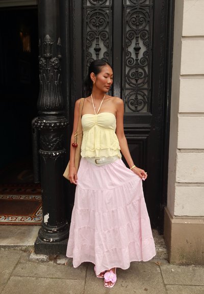Blusa amarilla con capas y tirantes finos, combinada con una falda maxi de capas en rosa claro, accessorios con un cinturón blanco y sandalias con flores.
