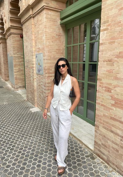 Mujer con un chaleco sin mangas y pantalones blancos caminando sobre una acera de baldosas hexagonales al lado de un edificio de ladrillos beige con una ventana de marco verde.