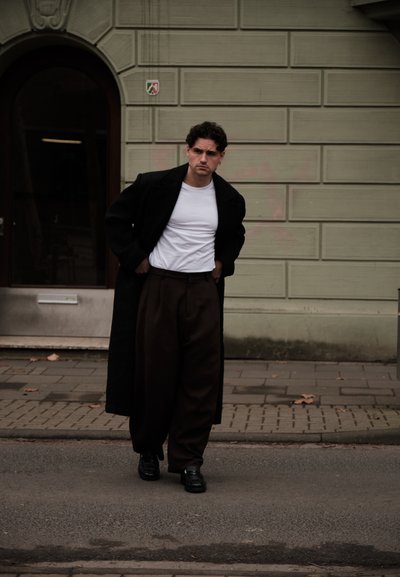 Hombre con un abrigo largo negro, camisa blanca y pantalones anchos oscuros de pie en una acera frente a un edificio de ladrillos beige.