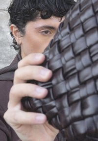 Jongere met krullend donker haar houdt een donkerbruine, gevlochten leren tas omhoog, die gedeeltelijk hun gezicht bedekt, tegen een neutrale achtergrond.