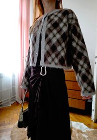 Άτομο που φοράει γκρίζο και μαύρο καρό καζάκα με κουμπιά πάνω από μαύρο ντύσιμο, κρατώντας μια τσάντα, στέκεται σε δωμάτιο με ξύλινο πάτωμα και κουρτίνες.