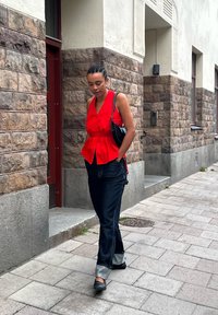 Raudona be rankovių palaidinė su peplum dizainu, tamsios džinsinės kelnės su apvadu, juoda petine rankinė, einanti akmeniniais šaligatviais.