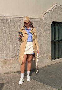 Manteau beige, chemise rayée bleu clair, mini-jupe blanche, collants à pois, baskets blanches et casquette beige. Tenant un smartphone.