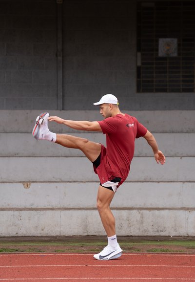 Atleta con ropa deportiva roja y gorra blanca estira la pierna hacia adelante en una pista de atletismo con gradas de concreto en el fondo.